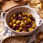 An image of a bowl of the warm marinated olives, sitting on a countertop surrounded by other ingredients in the background