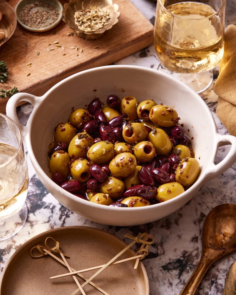 An image of a bowl of the warm marinated olives, sitting on a countertop surrounded by other ingredients in the background