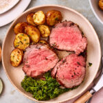 The sliced beef tenderloin, sitting on a plate next to crispy smashed potatoes and grilled broccolini