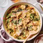 A big pot of the one-pan "Marry Me" chicken orzo, sitting on a countertop with a spoon resting inside of it