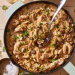 A pot full of the chicken marsala orzo, sitting on a countertop with a spoon resting in the pot