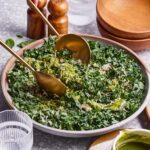 A bowl of the kale salad with lemon-basil dressing, sitting on a countertop with metal serving utensils resting in the bowl