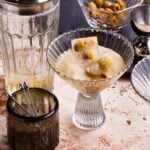 A glass of the quick and easy dirty martini, sitting on a countertop next to a cocktail shaker and a small bowl of olives