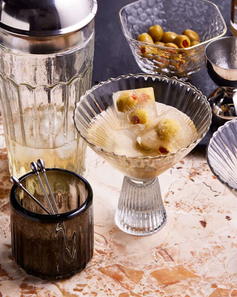 A glass of the quick and easy dirty martini, sitting on a countertop next to a cocktail shaker and a small bowl of olives