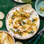 A bowl of the fried pickle dip, sitting on a countertop with a ruffled potato chip sitting in the bowl