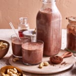 A jar of the easy chocolate cashew milk, sitting on a countertop, next to two smaller glasses of the cashew milk