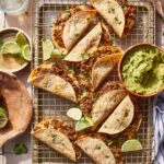 A tray of the crispy beef tacos, sitting on a wire cooling rack next to a bowl of guacamole