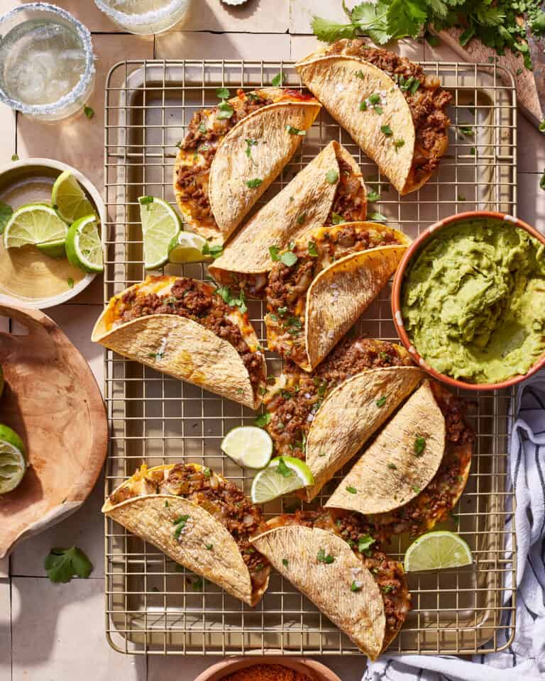A tray of the crispy beef tacos, sitting on a wire cooling rack next to a bowl of guacamole