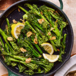 A pan of the crunchy garlic lemon broccolini, sitting on a countertop with lemon wedges and garlic