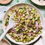 A bowl of the cucumber salad with crunchy quinoa, siting on a countertop with a serving spoon resting in the bowl