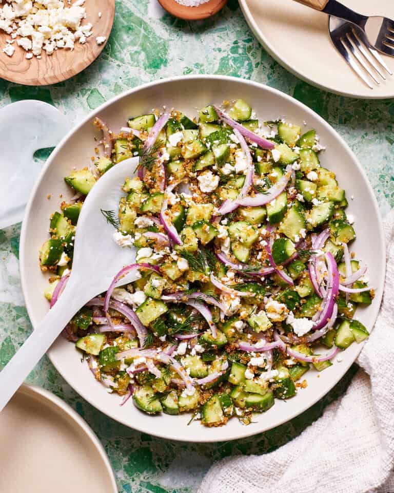 A bowl of the cucumber salad with crunchy quinoa, siting on a countertop with a serving spoon resting in the bowl