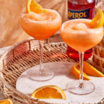 A glass of the easy frozen aperol spritz, sitting on a countertop topped with an orange slice and a bottle of aperol in the background