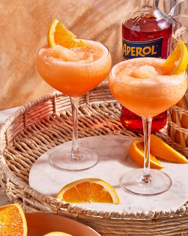 A glass of the easy frozen aperol spritz, sitting on a countertop topped with an orange slice and a bottle of aperol in the background