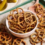 A bowl of seasoned pretzels sits on a parchment-lined baking sheet, with some pretzels scattered around. A wooden spatula rests nearby, and a plate with pickles is partially visible in the background.