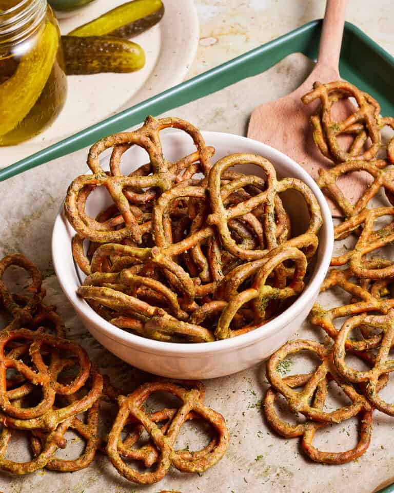 A bowl of seasoned pretzels sits on a parchment-lined baking sheet, with some pretzels scattered around. A wooden spatula rests nearby, and a plate with pickles is partially visible in the background.