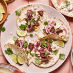 A plate of tacos filled with seasoned beef, corn, fresh cilantro, pickled red onions, and crumbled cheese. Lime wedges are placed around the tacos on the plate. Various bowls of ingredients and garnishes surround the plate on a pink tiled surface.
