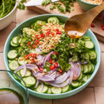 A vibrant salad in a light blue bowl features sliced cucumbers, red onion, chopped red chili peppers, cilantro, and peanuts. A spoon is held above, pouring dressing over the salad. Fresh herbs, a knife, and a partially cut lime are visible around the bowl.