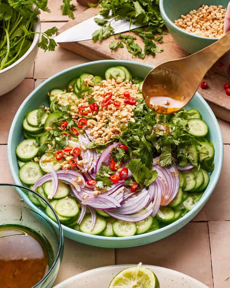 A vibrant salad in a light blue bowl features sliced cucumbers, red onion, chopped red chili peppers, cilantro, and peanuts. A spoon is held above, pouring dressing over the salad. Fresh herbs, a knife, and a partially cut lime are visible around the bowl.