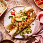 A plate of enchiladas topped with sliced avocado, cherry tomatoes, cilantro, and green chilies sits on a marble table. Nearby are a fork and knife, a dish of jalapeños, a glass of water, and a pink napkin.
