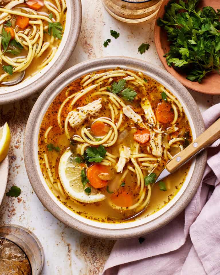 A bowl of chicken noodle soup with spaghetti, shredded chicken, carrot slices, and lemon wedge garnished with fresh parsley. There is a wooden spoon, a bunch of parsley in a bowl, a glass of beverage, a napkin, and another bowl of soup in the background.