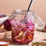 A jar of pickled red onions sits on a rustic wooden surface. The jar is filled with thinly sliced red onions soaking in brine and seasoned with herbs. A metal spoon is placed inside the jar. Nearby, there are bowls containing salt and dried herbs.