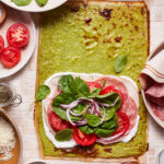 Top-down view of a flatbread topped with green pesto, slices of tomato, red onion, spinach leaves, and cured meats. Surrounding the flatbread are bowls of sliced tomatoes, spinach, spices, grated cheese, and cooking utensils on a light-colored surface.