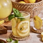 A refreshing glass of lemonade with lemon slices and basil leaves, placed on a woven coaster on a kitchen counter. A wicker basket filled with lemons, a pitcher of lemonade, and another glass with lemon slices are in the background. Basil leaves surround the glass.