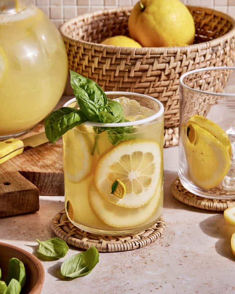 A refreshing glass of lemonade with lemon slices and basil leaves, placed on a woven coaster on a kitchen counter. A wicker basket filled with lemons, a pitcher of lemonade, and another glass with lemon slices are in the background. Basil leaves surround the glass.