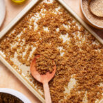 A golden baking tray filled with cooked quinoa, with a wooden spoon resting on top. Surrounding the tray are various bowls containing raw quinoa, oil, and other ingredients on a light-colored surface.