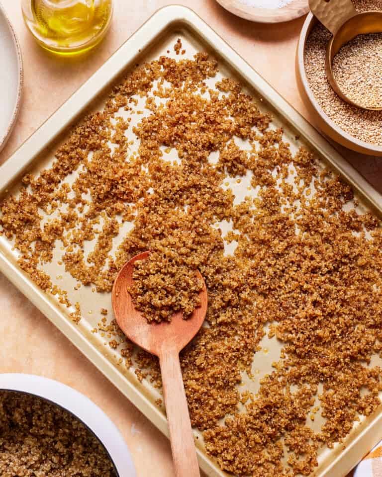 A golden baking tray filled with cooked quinoa, with a wooden spoon resting on top. Surrounding the tray are various bowls containing raw quinoa, oil, and other ingredients on a light-colored surface.