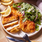 A plate with crispy breaded chicken cutlet partially sliced, accompanied by a fresh green salad with red onions and grated cheese. A lemon wedge is placed on the side. A fork and knife rest on the plate, and a drink is in the background.