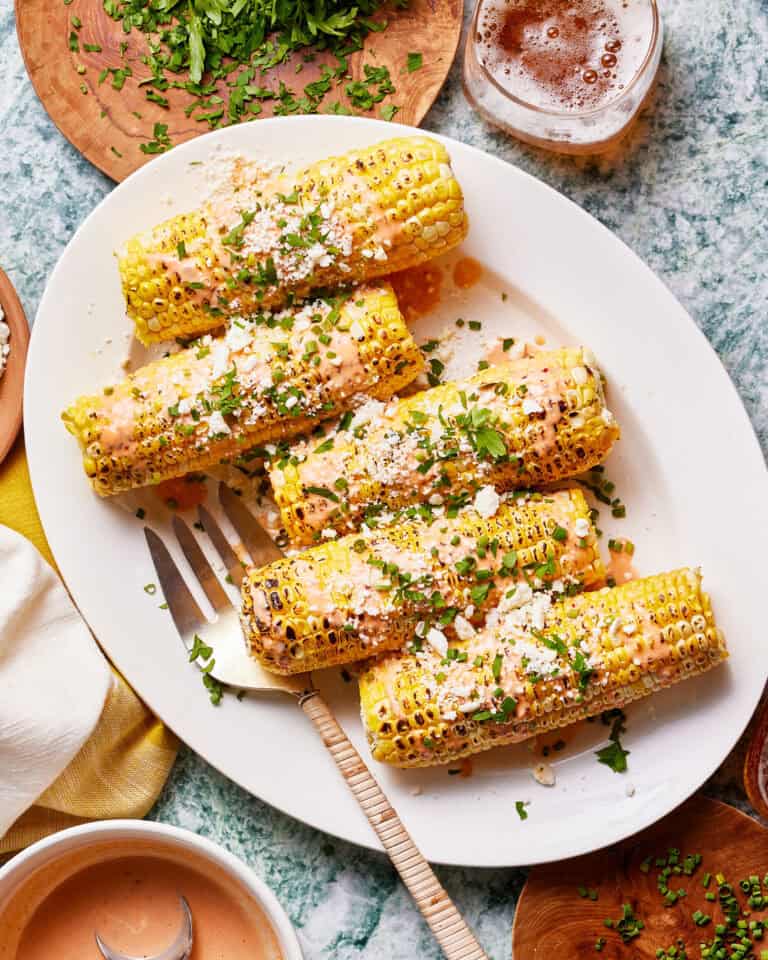 A plate with five grilled corn on the cob pieces topped with creamy sauce, crumbled cheese, and chopped herbs. A fork rests on the plate, and the background shows a glass of drink, chopped herbs, and a bowl of sauce. A napkin lies beside the plate.