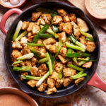 A skillet filled with stir-fried tofu and green onions sits on a marble countertop. Surrounding the skillet are various bowls containing ingredients like garlic cloves, white pepper, and sesame seeds. The vibrant green onions contrast with the golden-brown tofu.