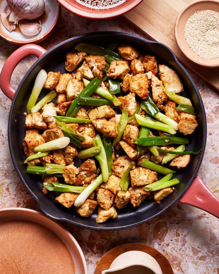A skillet filled with stir-fried tofu and green onions sits on a marble countertop. Surrounding the skillet are various bowls containing ingredients like garlic cloves, white pepper, and sesame seeds. The vibrant green onions contrast with the golden-brown tofu.