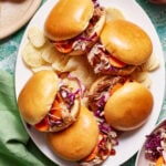 An overhead view of a plate of pulled pork sandwiches with coleslaw and BBQ sauce in soft buns. The sandwiches are surrounded by plain potato chips. A green napkin and part of another plate are visible in the background on a green marbled surface.
