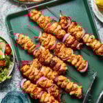 A green tray displays several skewers of grilled chicken and red onions. Tongs lie on the tray next to the skewers. In the background, there is a salad bowl with tomatoes and greens, a partially visible stack of pita bread, and glasses of water.