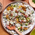 A plated dish of creamy potato salad garnished with crispy bacon bits, diced cucumbers, and chopped herbs. A wooden spoon rests in the salad, and the plate is placed on an orange cloth with some green plates and small dishes nearby.