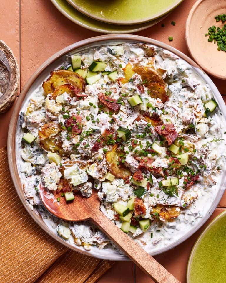 A plated dish of creamy potato salad garnished with crispy bacon bits, diced cucumbers, and chopped herbs. A wooden spoon rests in the salad, and the plate is placed on an orange cloth with some green plates and small dishes nearby.