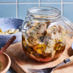 A glass jar filled with marinated artichokes is placed on a wooden cutting board. The jar is surrounded by a small wooden spoon with herbs, a bowl with artichoke pieces, and a fork resting on the board. The background features a light blue tiled wall.
