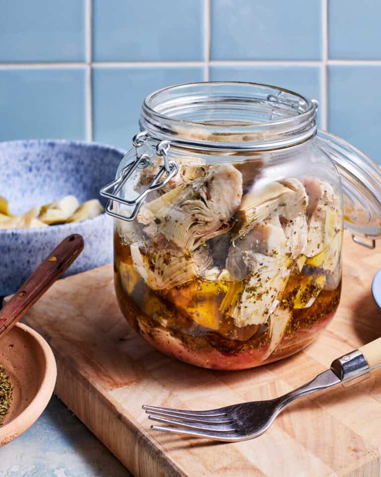 A glass jar filled with marinated artichokes is placed on a wooden cutting board. The jar is surrounded by a small wooden spoon with herbs, a bowl with artichoke pieces, and a fork resting on the board. The background features a light blue tiled wall.