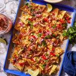 A colorful sheet pan of loaded nachos topped with shredded meat, melted cheese, diced tomatoes, corn kernels, sliced red onions, chopped cilantro, and lime wedges. The nachos sit on a parchment paper-lined blue tray set on a marble surface.