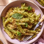 A bowl of pasta topped with vibrant green pesto sauce, garnished with fresh basil leaves and a sprinkle of breadcrumbs. The dish is served in a pink bowl with a fork on the side. There are additional pasta bowls and a cloth napkin in the background.