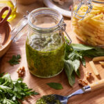 A jar of pesto sauce sits on a wooden cutting board, surrounded by fresh basil leaves, parsley, garlic, walnuts, and a spoon with pesto on it. In the background, there's an open glass jar of uncooked penne pasta and a bowl of garlic cloves.