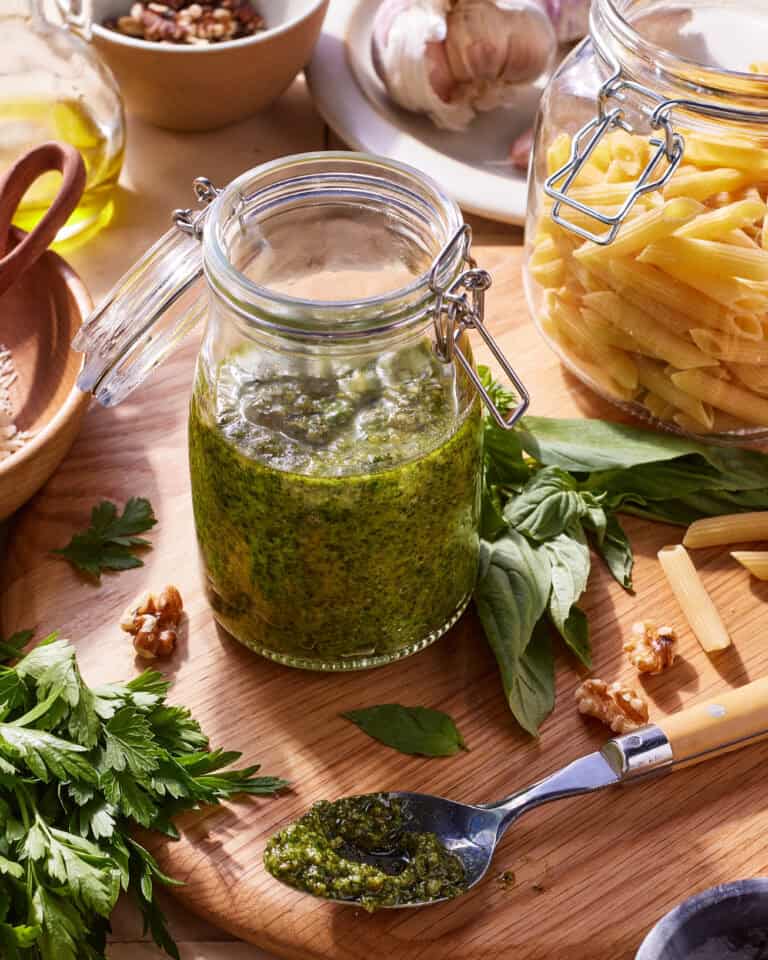 A jar of pesto sauce sits on a wooden cutting board, surrounded by fresh basil leaves, parsley, garlic, walnuts, and a spoon with pesto on it. In the background, there's an open glass jar of uncooked penne pasta and a bowl of garlic cloves.