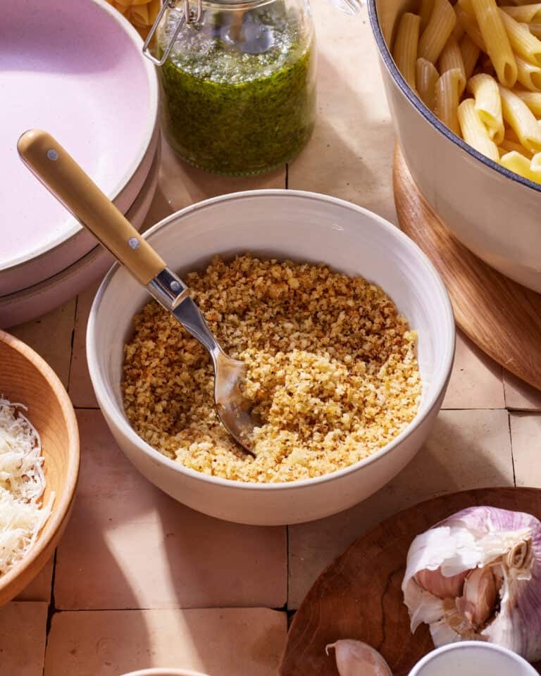 A bowl filled with breadcrumb mixture, placed next to a spoon, sits on a tiled surface. Surrounding it are containers of pesto sauce, uncooked penne pasta, grated cheese, and a head of garlic. The ingredients suggest preparation for a meal.