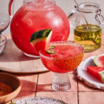 A refreshing pink cocktail garnished with a watermelon slice, chile powder, and a jalapeño slice served in a glass with a chile powder rim. A large pitcher of the same drink and a jar of infused liquid are in the background along with a cutting board with watermelon slices.