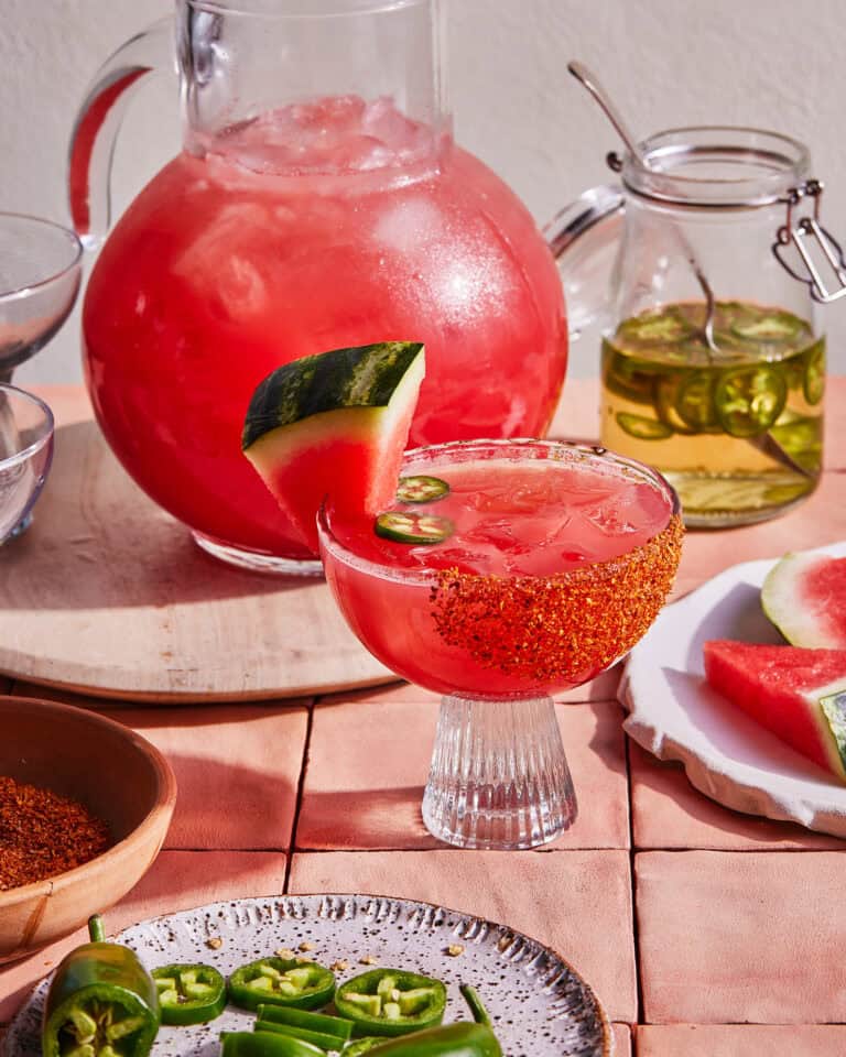 A refreshing pink cocktail garnished with a watermelon slice, chile powder, and a jalapeño slice served in a glass with a chile powder rim. A large pitcher of the same drink and a jar of infused liquid are in the background along with a cutting board with watermelon slices.