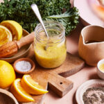 A jar of green vinaigrette dressing with a spoon inside sits on a wooden board. Surrounding it are fresh kale leaves, lemon halves, lemon slices, a small bowl of salt, a small bowl of ground pepper, a jug of oil, and a plate. A cut shallot is also visible.