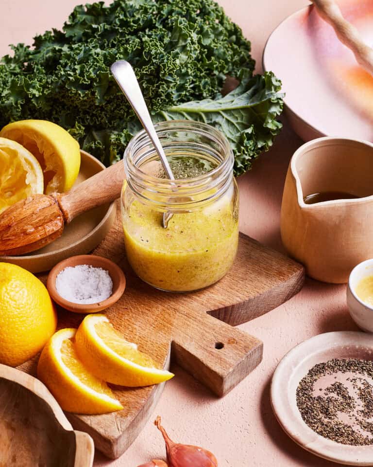 A jar of green vinaigrette dressing with a spoon inside sits on a wooden board. Surrounding it are fresh kale leaves, lemon halves, lemon slices, a small bowl of salt, a small bowl of ground pepper, a jug of oil, and a plate. A cut shallot is also visible.