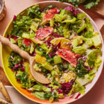 A vibrant salad in a white bowl topped with various fresh greens, avocado, segments of citrus fruits like blood orange, and sprinkled with sunflower seeds. A wooden spoon rests in the salad, ready to serve. Pieces of kale and sliced radicchio are visible.